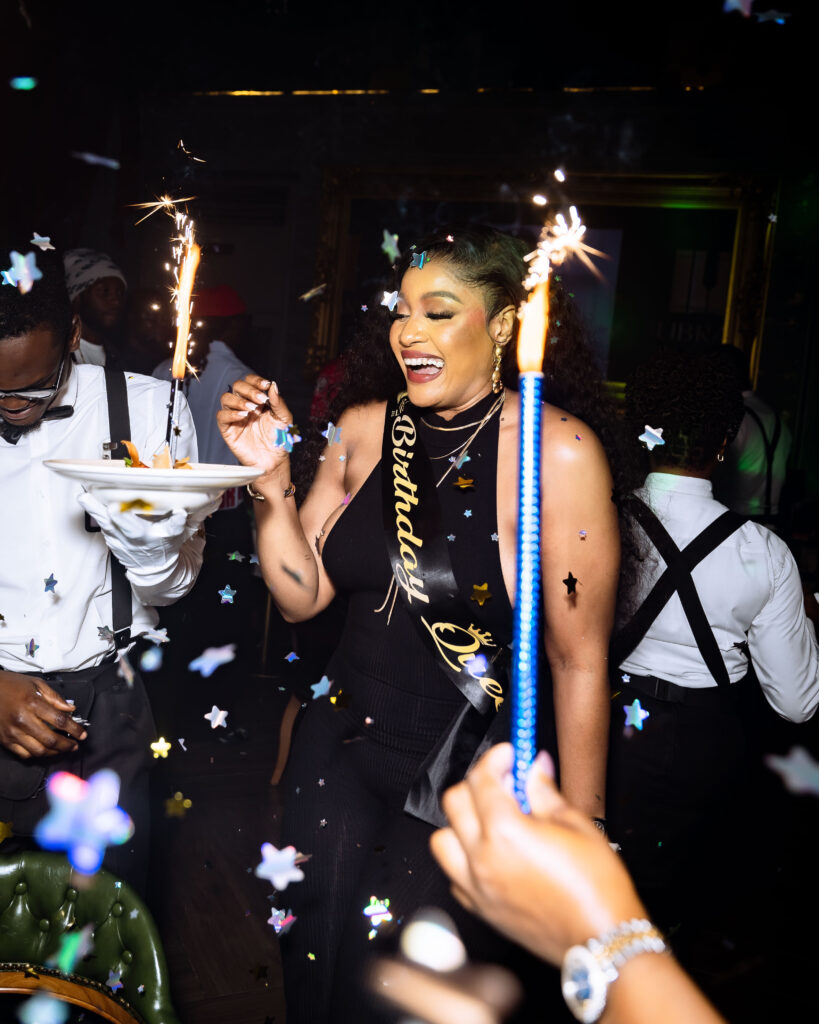 A Scholar at The Library Lagos laughing joyfully as two waiters stand beside her, popping confetti while a dessert plate sparkles with light at the center of the celebration