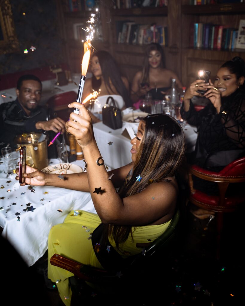 A group of friends at The Library Lagos celebrating a birthday, raising their hands and phones to capture the celebrant as confetti pops and sparklers light up the air, their faces glowing with admiration and excitement